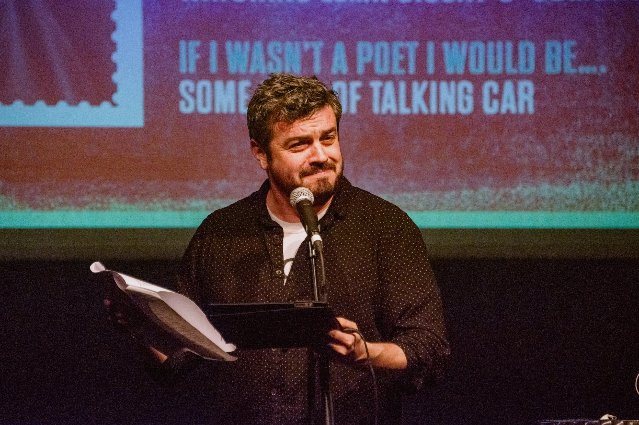 Man In a spotted shirt reading to audience on stage