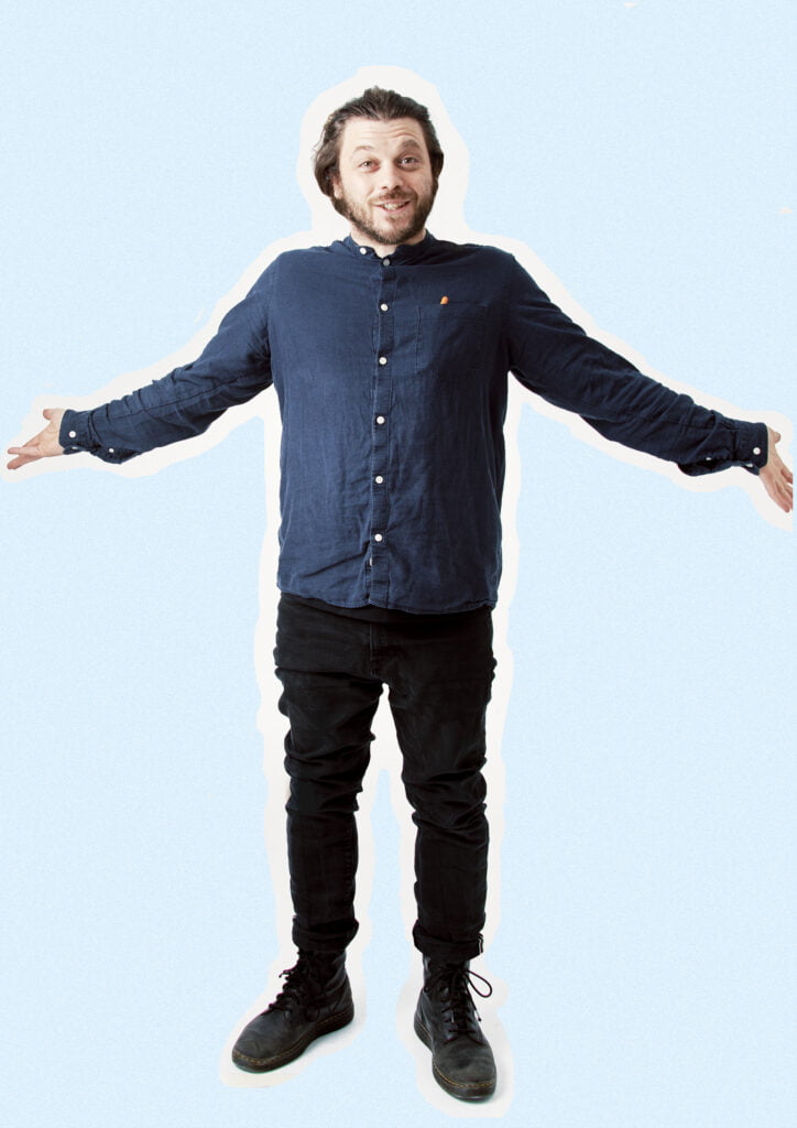 An image of poet Adam Kammerling. He wears a blue shirt and black trousers, he wears black trainers. His arms are outstretched to each side of his body as he smiles at the camera. He has long shoulder length brown hair and a brown beard.
