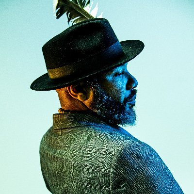 A man in a feather hat and tweet jacket looks sideways on