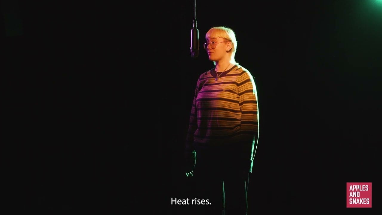 Screenshot from a video of Cherry speaking into a microphone in a dark room with yellow and pink lighting