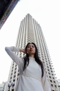 A woman with long dark hair wearing a high necked, long sleeve white dress puts her arm behind her head, standing in front of a tall tower block