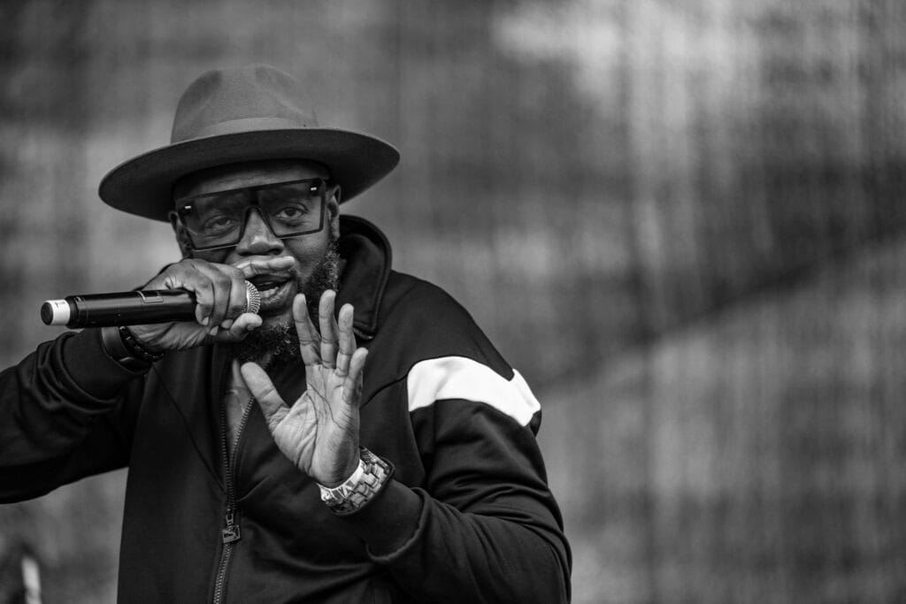 BREIS performs wearing glasses and a wide-brimmed hat speaks into a microphone with expressive hand gestures, captured in black and white.