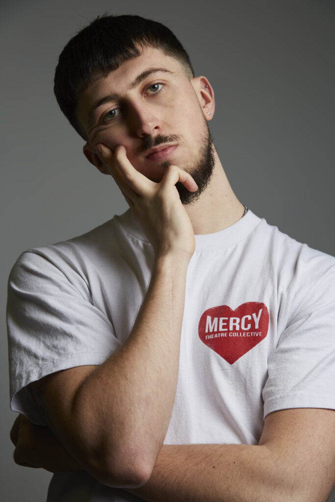A photo of poet David Woolcott. David has short black hair and a short beard. He wears a white t-shirt with a red heart printed on the right hand side. Inside the heart it says Mercy Theatre Collective. He holds his hand up to his face leaning his head into his hand. 