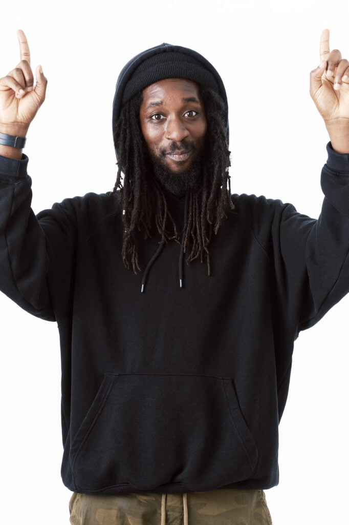An image of poet Ehireme Omoaka. He has long black hair and wears a black hoodie and Green trousers. He points his to hands towards the sky and smiles at the camera.