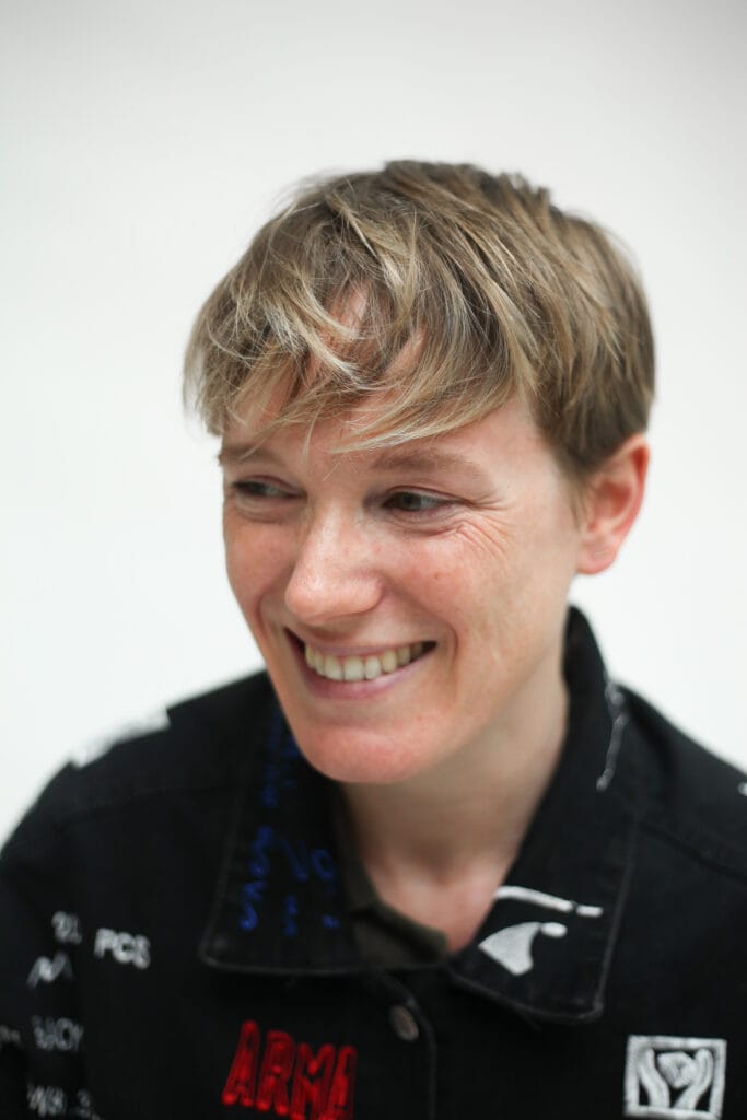 A non-binary person with short side swept blond hair, smiling and wearing. a black shirt with small motifs on.