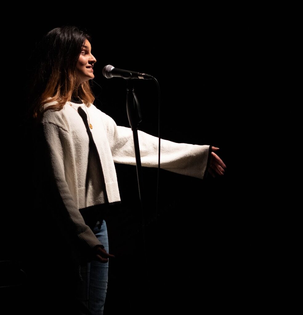 An image of poet Noir Iman. She has shoulder length brown hair and wears a white jumper and blue jeans. She stands in front of a microphone and is performing a poem.
