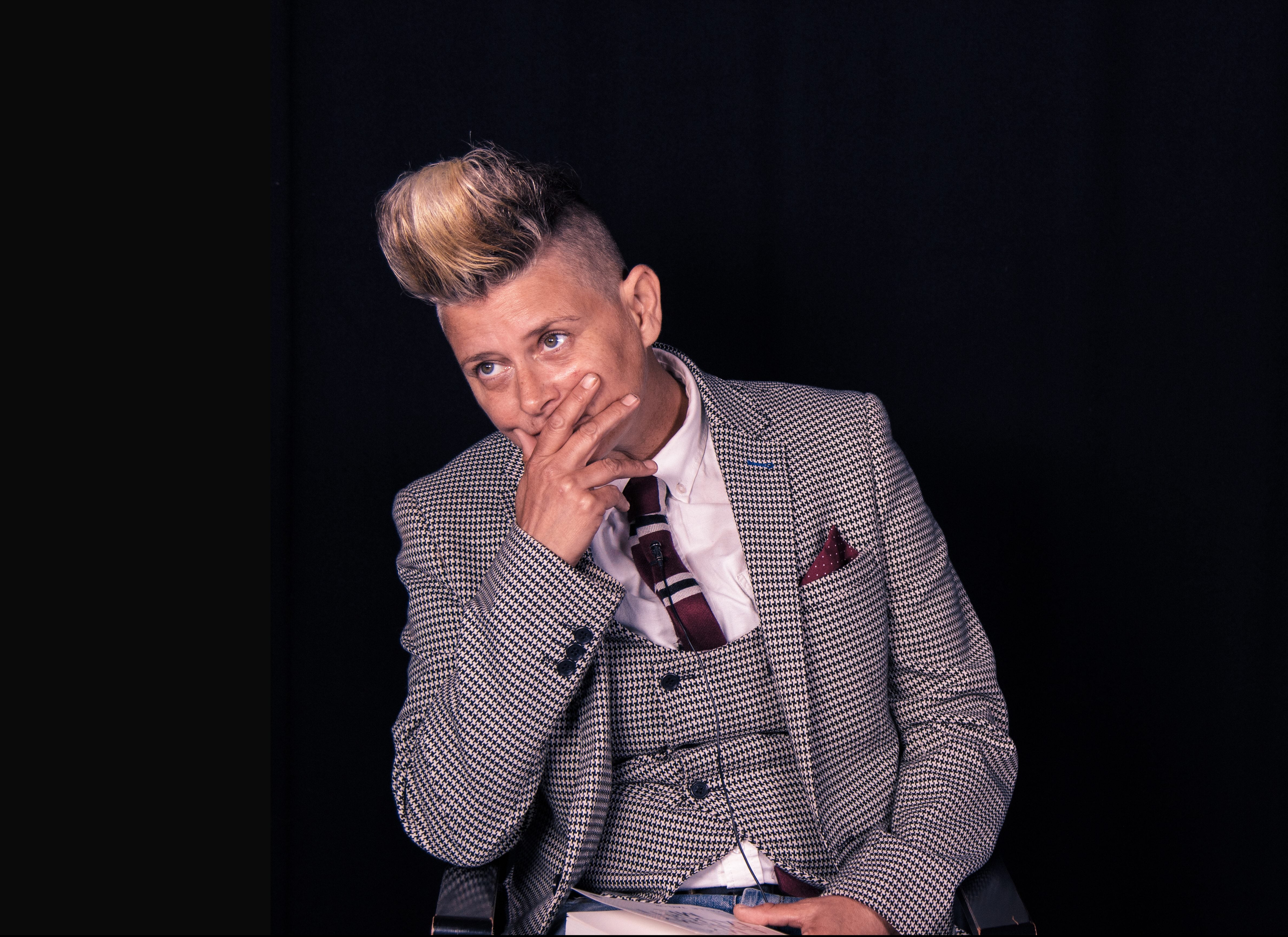 Picture of Joelle Taylor wearing a waistcoat, jacket and tie. Joelle is looking to the left and holding her mouth.