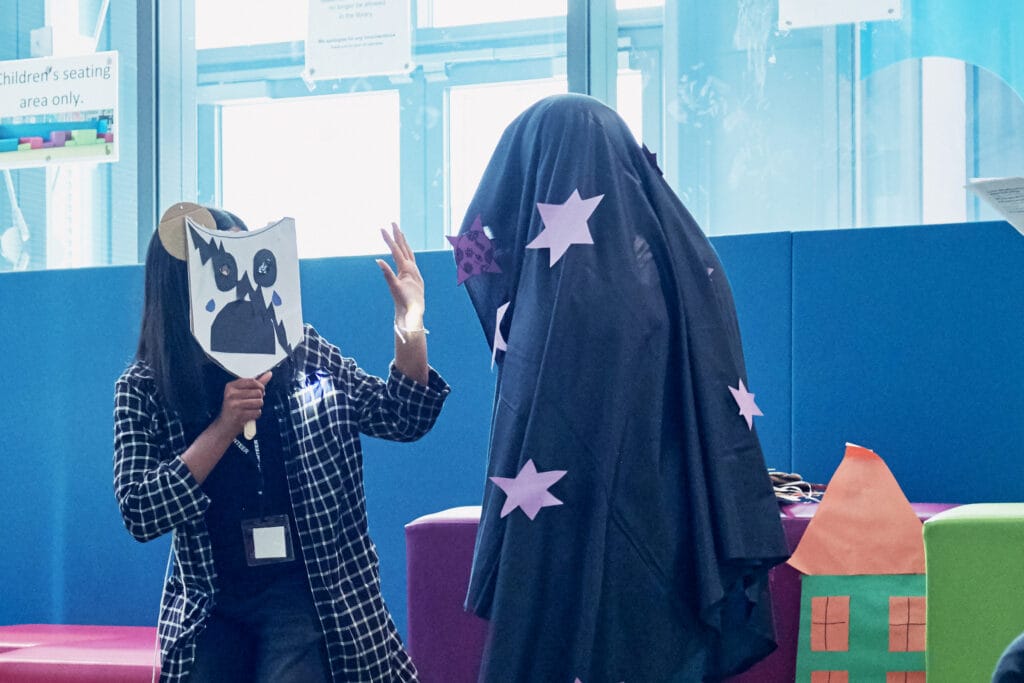 Two people perform a playful scene in a brightly lit library setting. On the left, a person in a checkered shirt holds up a handmade mask featuring a cartoonish, wide-eyed, shocked face with tear shapes and horns. Their other hand is raised mid-gesture. On the right, another person is fully draped in a dark cloth decorated with large purple paper stars, appearing like a costumed character. Colorful paper props, including a cut-out house, are visible in the background. The setting suggests a children's storytelling or drama activity.