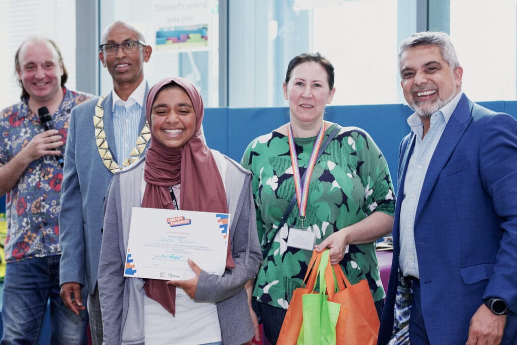 A joyful young woman wearing a maroon headscarf smiles broadly while holding a “Library Takeover” certificate of recognition. She is surrounded by four adults who are also smiling and celebrating the moment. One man, wearing a floral shirt, holds a microphone. Another, wearing a ceremonial mayoral chain, stands beside him. A woman in a green patterned dress with a lanyard and two colorful tote bags stands to the right of the awardee. On the far right, a man in a blue suit jacket smiles warmly. They appear to be at an indoor event, likely a celebration or award ceremony.