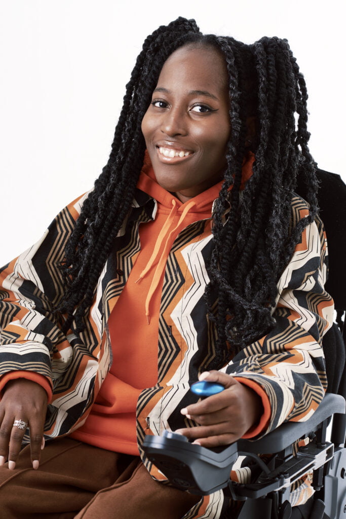 An image of Poet Miss Jacqui, She has long black hair and wears an orange hoodie, she also has a brown, orange and white coat on, she wears brown trousers. She smiles at the camera, her left hand rests on the blue controls of her wheelchair.