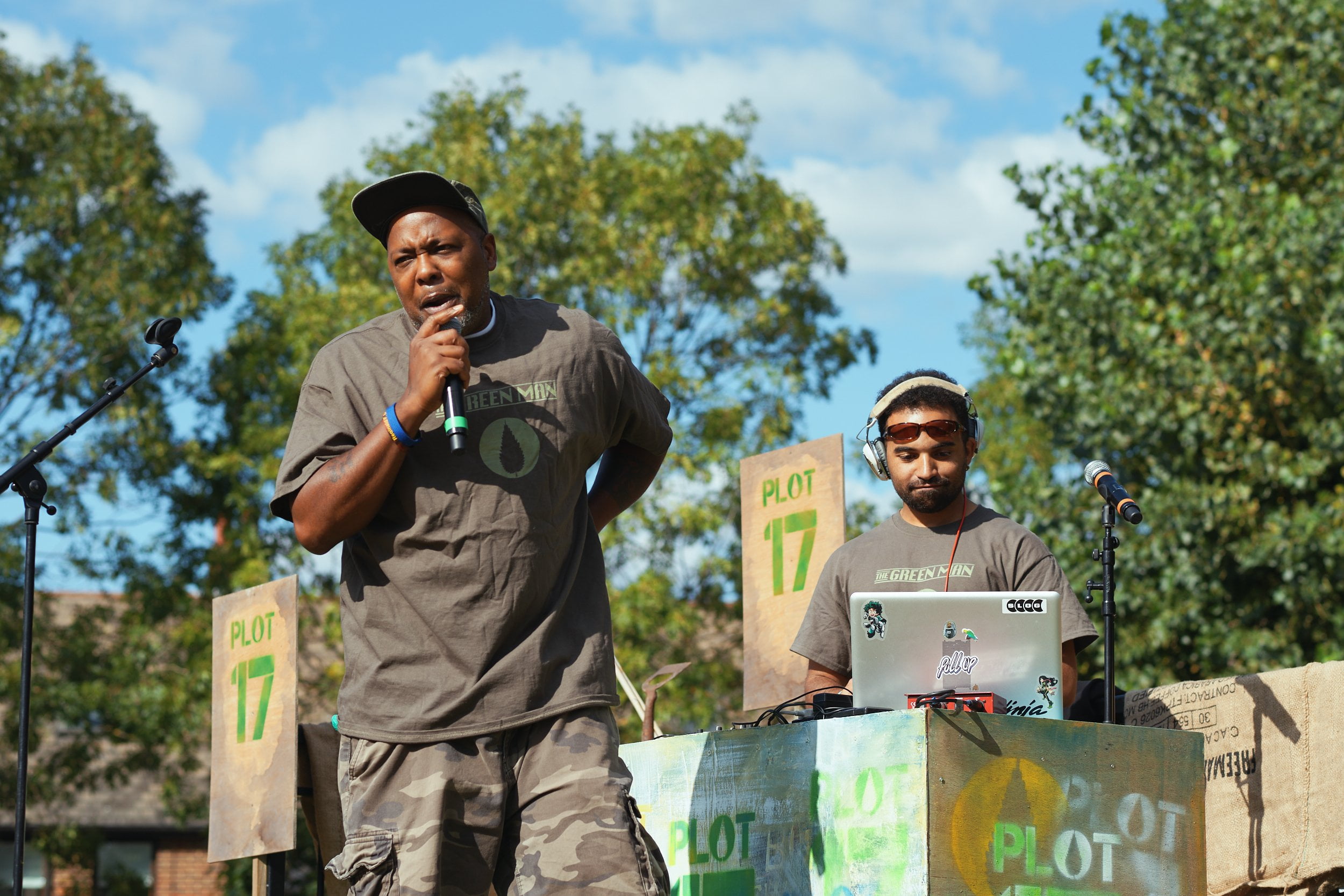 Two men on stage - one with a laptop and headphones and one with a cap speaking into a microphone