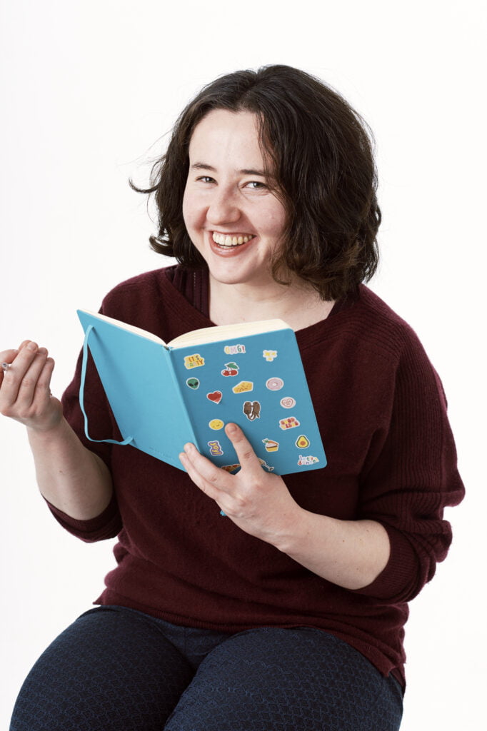 An image of poet Rachel Lewis. She has shoulder length brown hair. She smiles at the camera and holds a blue book covered in stickers. She wears a brown jumper and black trousers.