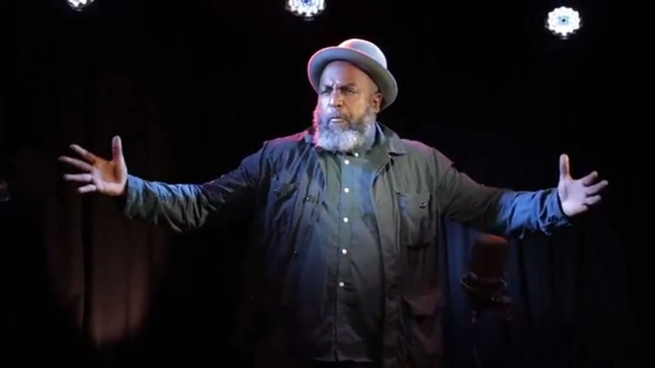 Image of a man with a grey hat and beard with arms outstretched