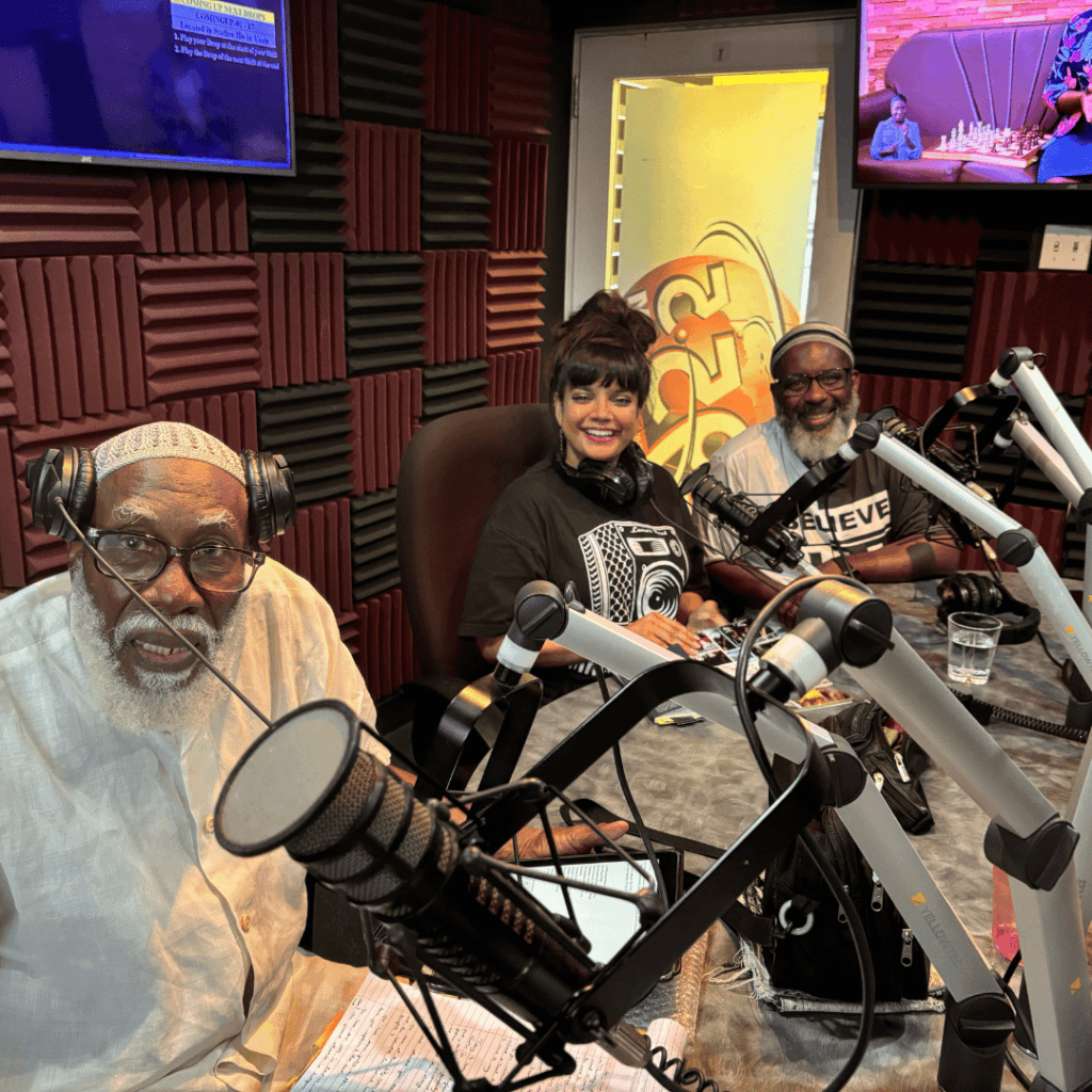 The setting in this photo is a radio studio. Three people are sat at a desk wearing headphones and have radio mics in front of them on the desk. They all look and smile at the camera. 