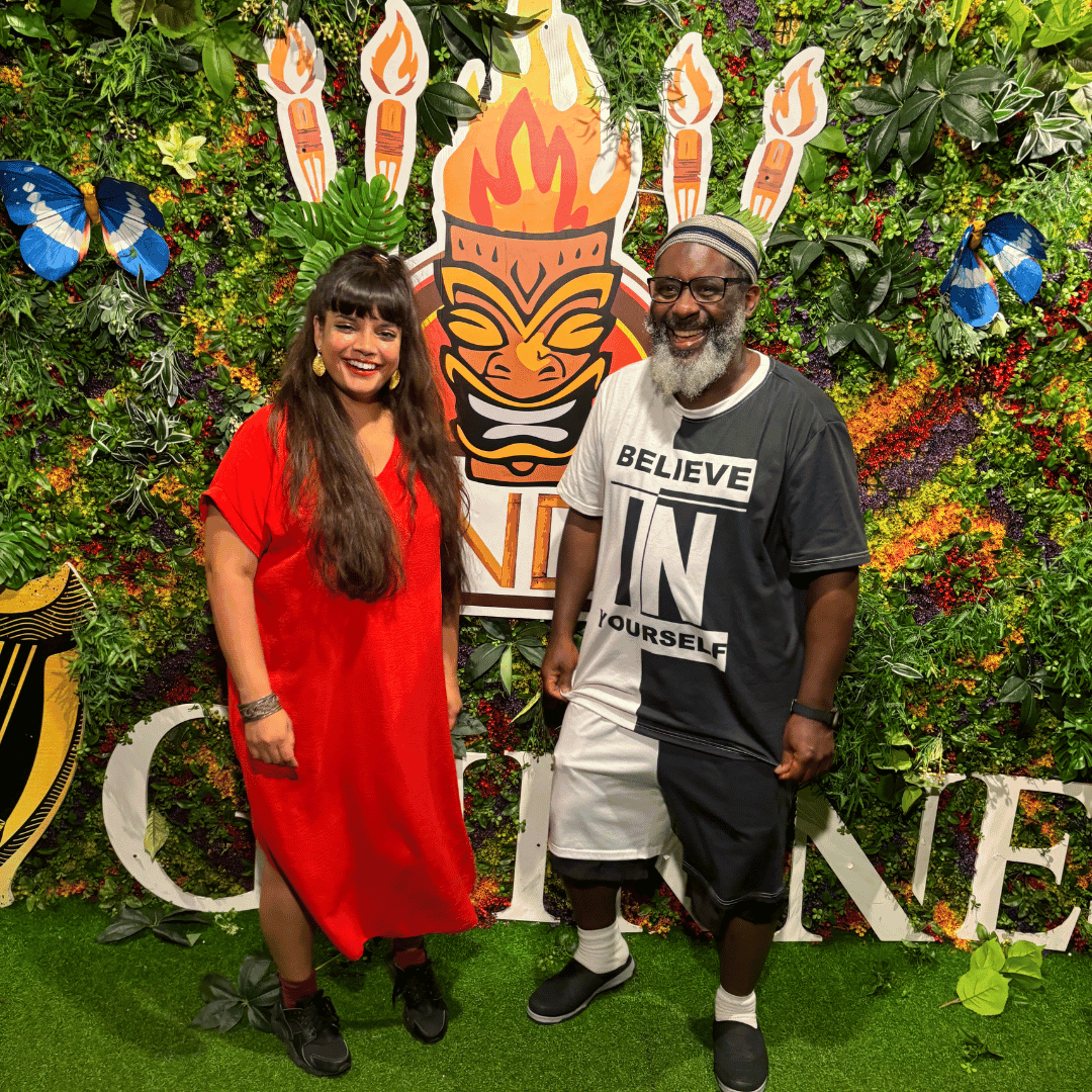 2 people stand in front of a fire mural, on the left a woman with long black hair and wearing a red dress. on the right a man, wearing a black and white t-shirt and black and white shorts. They both smile at the camera
