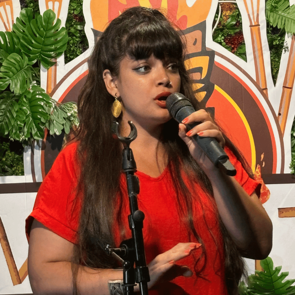 Poet Shareefa Energy stands holding a microphone, she wears a red t-shirt and stand in front of a green plant. She has long brown hair.