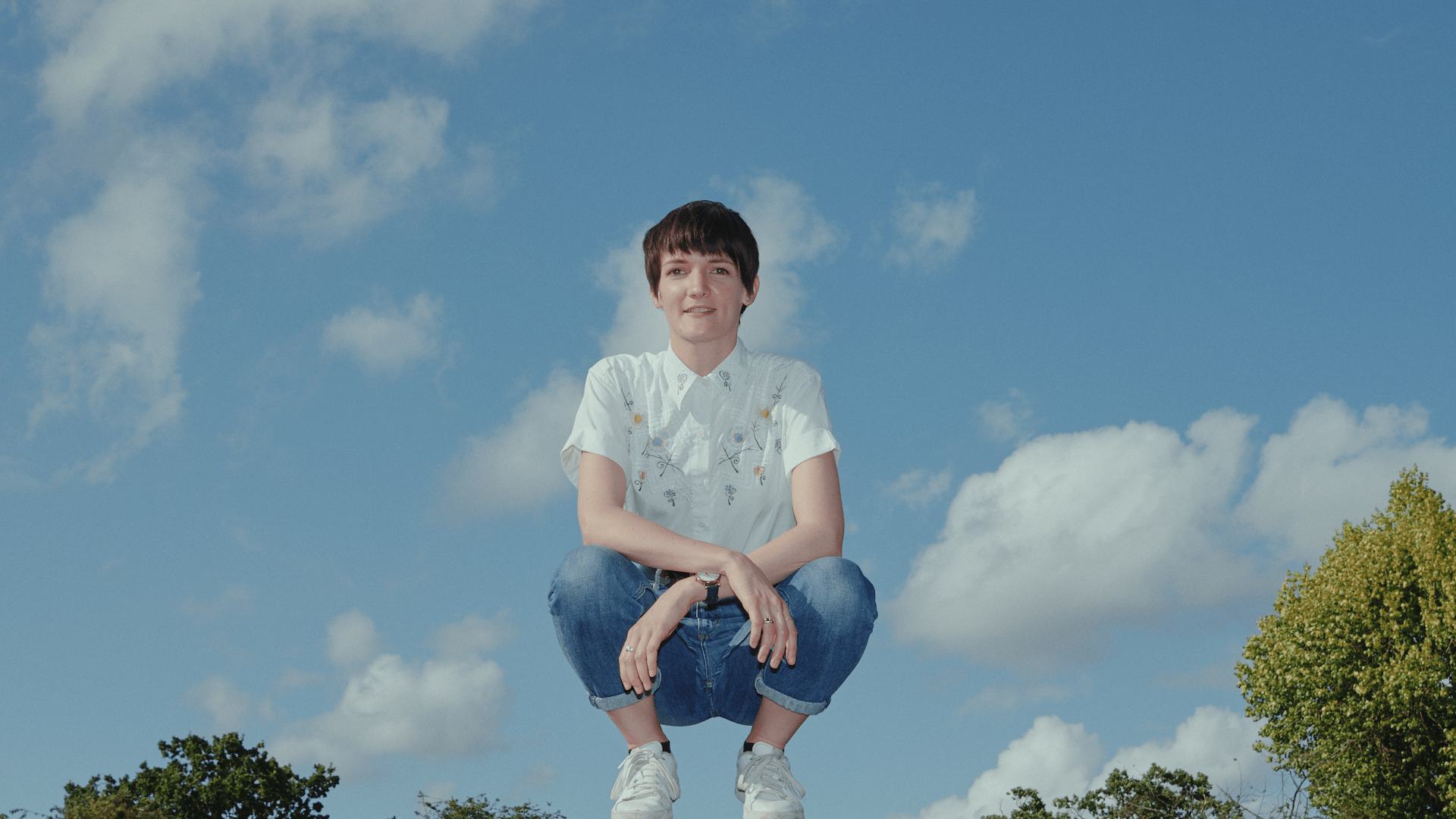 A woman with short hair, white shirt and blue jeans squats on a brick wall against a blue sky