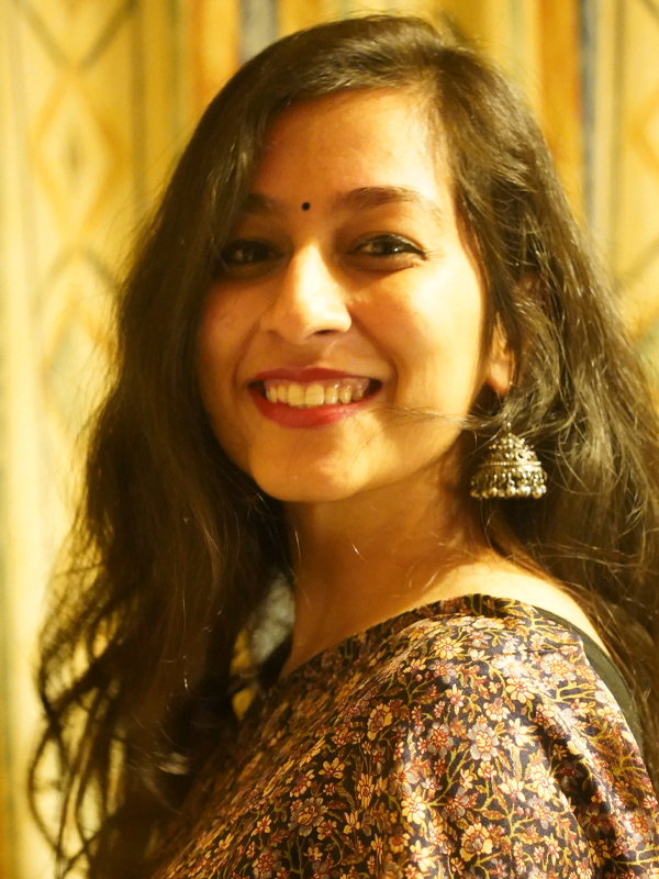 Bhumika wears bell shaped earrings with a tight floral pattered dress, hair is dark, wavy and long