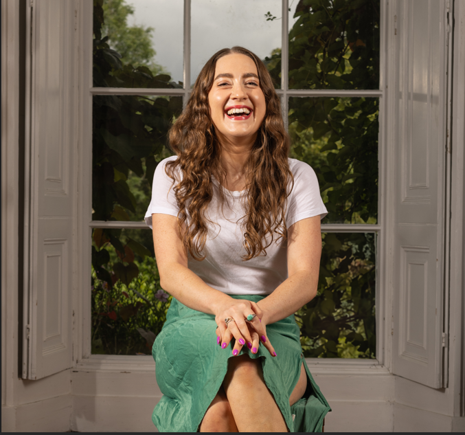 An image of poet Beth Kelly. She sits in front of a window and crosses her legs, her hands are placed on her knees. She wears a green skirt and white t-shirt. She smiles towards the camera. She has long, brown, curly hair and pink fingernails. She has a green ring on her Index finer.