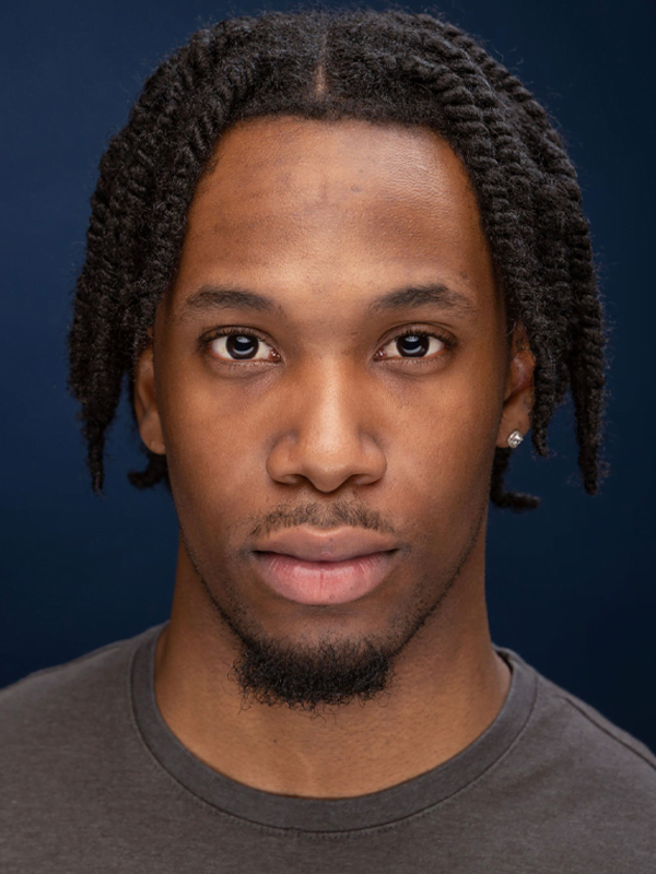 Blaize has twisted braids that are chin length, a gem stone earring and dark grey top against a dark blue background