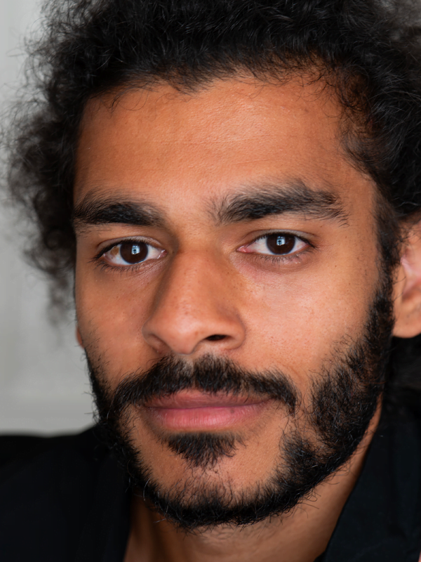 Dan has dark curly hair pulled up, with a dark trimmed beard, wearing a dark collared top