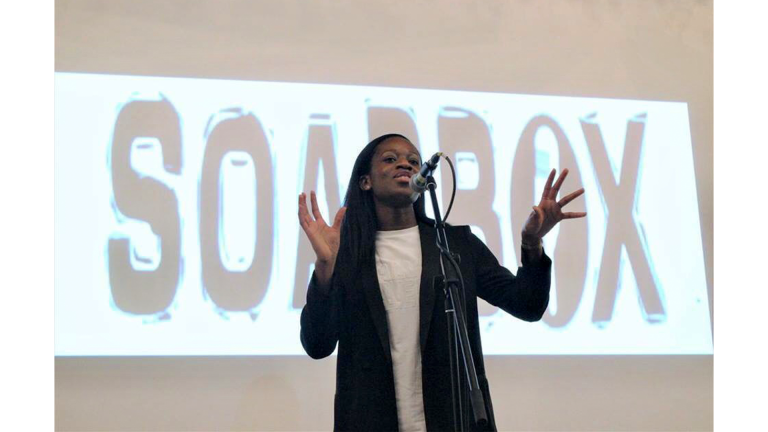 A woman in a black jacket and white shirt on stage, talking into a mic