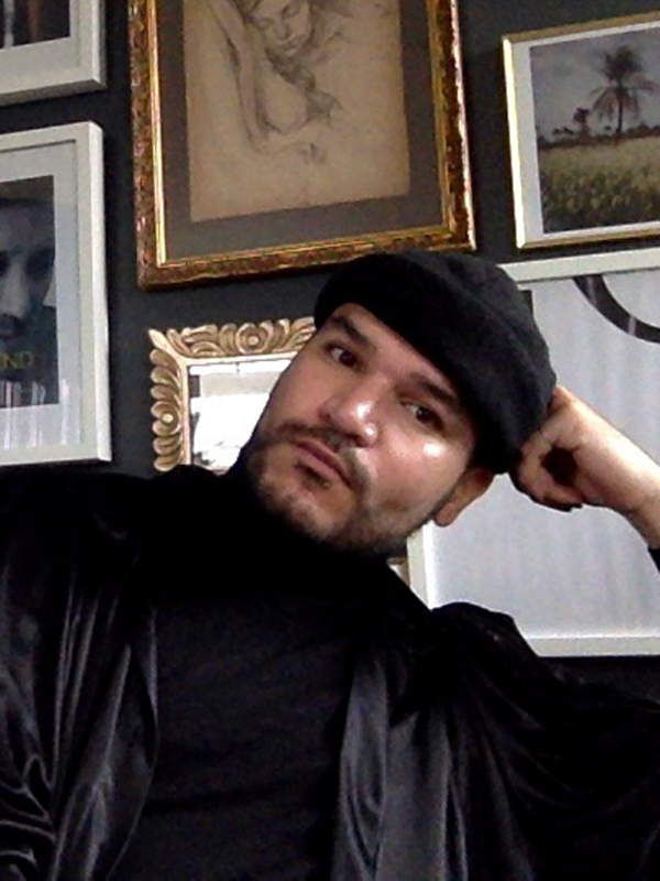 Gaetan has a black jacket, t-shirt and woollen hair, resting his arm against his head. Various portraits and framed art in the background