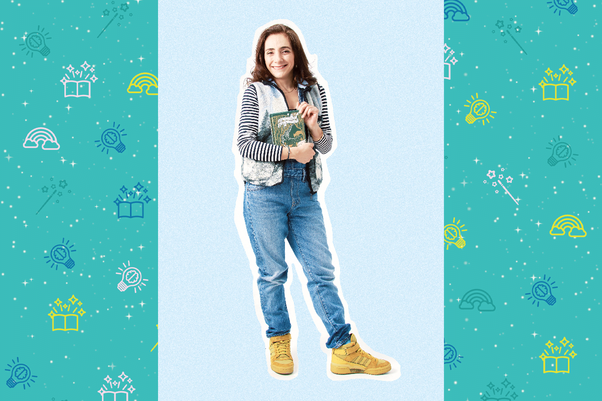 An image if artist Gunes Given, she holds a green book. She has brown shoulder length hair and wears a blue sleeveless cardigan with a black and white stripy top underneath. She wears blue jeans and yellow sneakers. She smiles at the camera.
