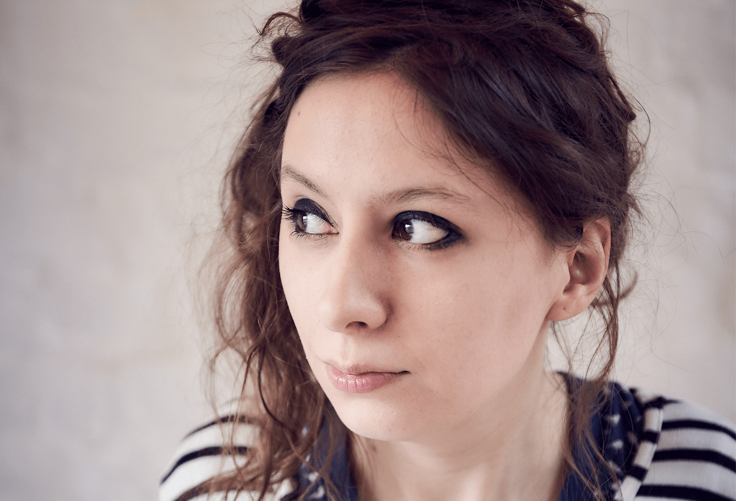 A woman with dark eye make up and loosely pulled up dark brown hair, looking sheepishly to the left