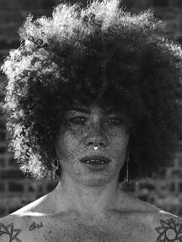 Holly has a large afrro type hair and lots of freckles. with sun like tattoos on her shoulders and a nose piercing.