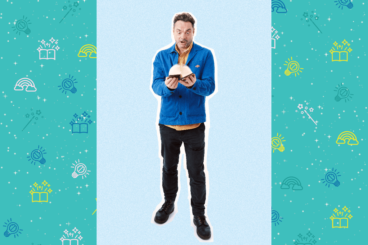 An image of poet Justin Coe, he has short brown hair and wears a blue jacket with an organ shirt underneath. He holds a book in front of him and looks at it. He is wearing black trousers and black shoes.