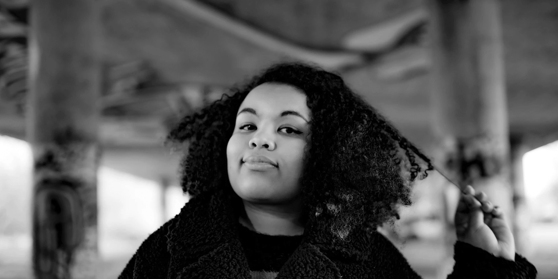 A woman with shoulder length tight curly dark hair runs her hand through one side and smiles
