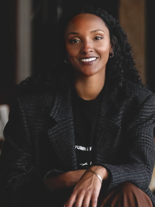 Mariah smiles, wearing her dark curly hair down. She is wearing a wool grey and black blazer, and black tshirt with a silver bracelette