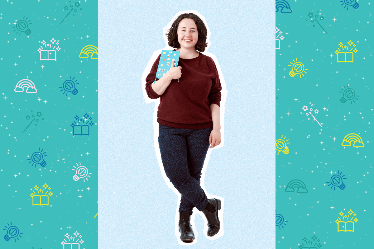 An image of poet Rachel Lewis. She has shoulder length brown hair. She smiles at the camera and holds a blue book covered in stickers. She wears a brown jumper and black trousers.