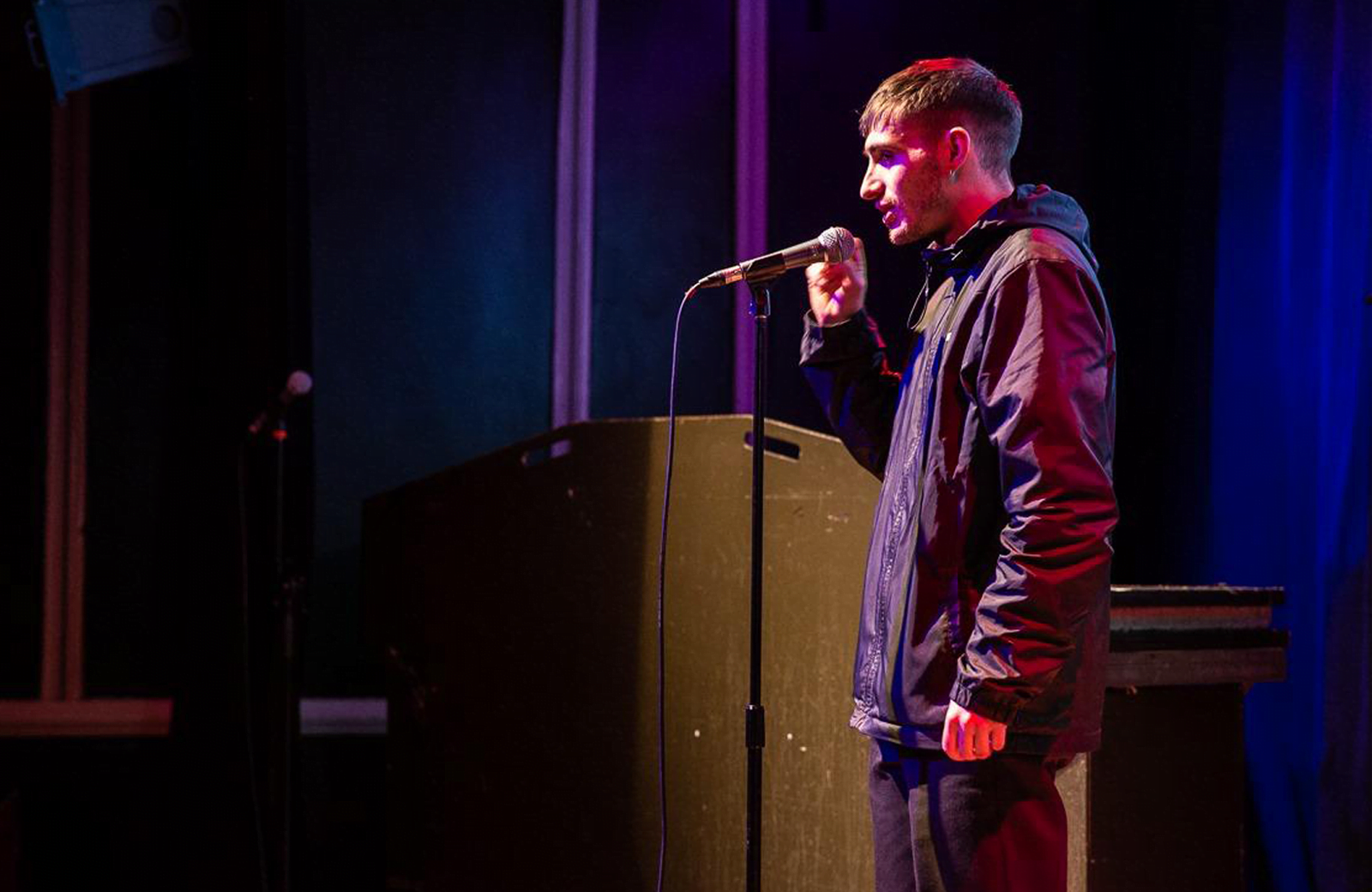A young man standing on stage, speaking into a mic, wearing a blue outdoors rain mac