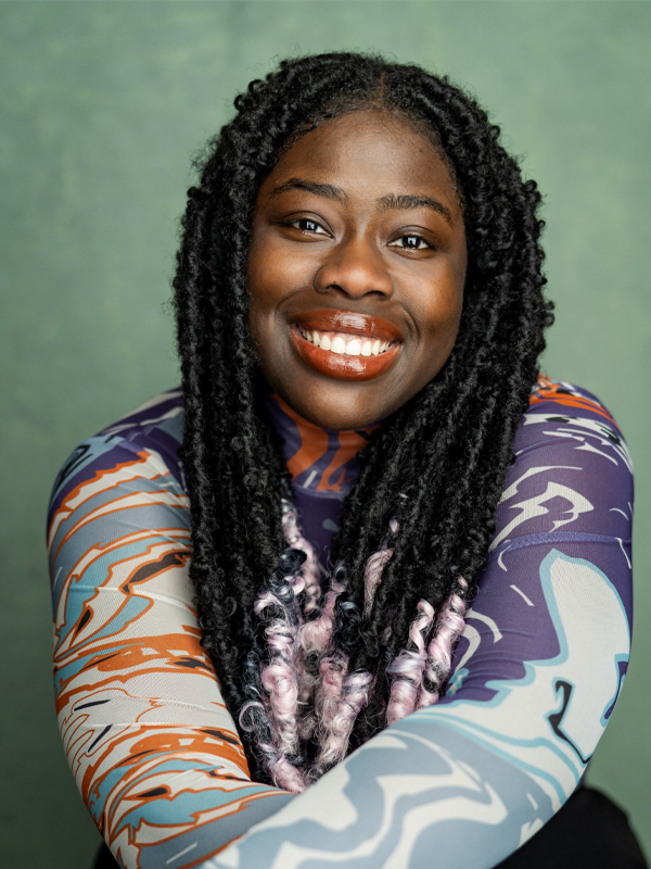 Seun wearing a blue, purple and orange swirly long sleeved top, long braided hair with pink tips. She is smiling and wearing orange lipgloss