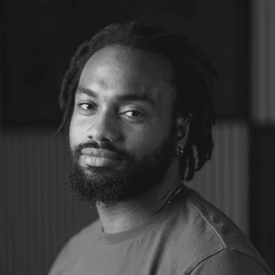 A black adult male with a beard and short locks, wearing a plain t-shirt